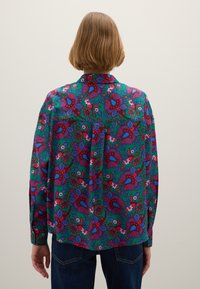 Person mit kurzen, hellbraunen Haaren, die eine grüne Bluse mit einem roten und blauen floralen Paisleymuster trägt, steht vor einem einfarbigen Hintergrund.