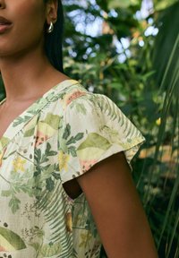 Bluse med blomsterprint med korte puffærmer, lysegrøn og gul stof med blade og blomster, der fremhæver en naturlig, tekstureret finish.