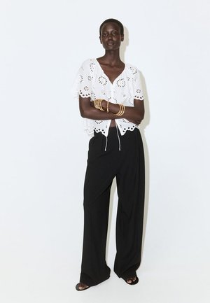 Femme debout, bras croisés, portant une blouse blanche à œillets, un pantalon noir à jambes larges, des sandales noires et plusieurs bracelets dorés au bras droit.