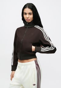 Καφέ κοντό μπουφάν με φερμουάρ και ψηλό γιακά, με λευκές ρίγες στα μανίκια. Διαθέτει μικρό λογότυπο και συνοδεύεται από λευκό παντελόνι με μπορντό λεπτομέρειες.