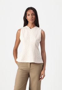 Lys beige ermeløs topp med et teksturert blomstermønster, med V-hals og en tettsittende silhuett, kombinert med brune bukser.
