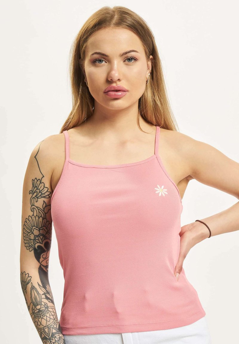 Jonge vrouw met lang blond haar, draagt een roze tanktop met kleine witte bloemdetails, poseert met een hand op haar heup en toont bloem tattoos op haar armen.