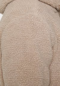 Tela de forro polar beige con una superficie texturizada y suave, que presenta una apariencia esponjosa y un patrón ligeramente irregular.