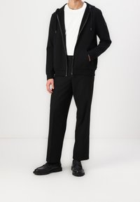 Man som bär en svart zip-up hoodie, vit t-shirt, svarta raka byxor och svarta läderskor, står mot en vit bakgrund.