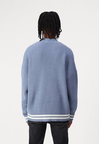 Blå stickad tröja med ribbat mönster, oversized silhuett och randiga vita detaljer vid fåll och ärmslut. Lätt och avslappnad design.