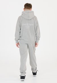 Grå hoodie med logotypen "Club de Sport Les Arcs" på baksidan, ihop med matchande sweatpants. Materialet ser mjukt ut; avslappnad passform. Svarta sneakers bärs.
