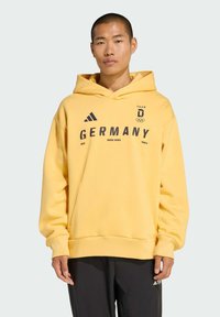 Gelber Kapuzenpullover mit einer vorderen Kängurutasche, versehen mit dunklem Text und Logos: "DEUTSCHLAND", "WINTERSPORT" und Olympische Symbole. Baumwollmischstoff.