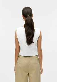 Weißes ärmelloses Top mit taillierter Passform, das eine glatte Textur und einen abgerundeten hinteren Saum präsentiert, kombiniert mit beigen hoch geschnittenen Hosen.