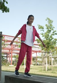 Różowy dres sportowy z kapturem na zamek i elastycznymi spodniami. Biała koszulka pod spodem z małym czarnym logo. Czarne sneakersy. Plener.