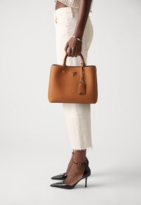 Brown leather handbag with a structured design, top handles, and a removable tag. Paired with white cropped pants and black pointed-toe heels.