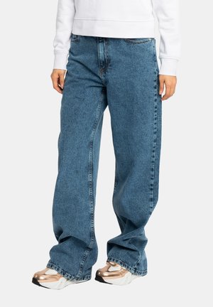 Personne portant un jean en denim bleu ample, un sweat-shirt blanc et des baskets blanches avec des accents dorés métalliques, debout sur un fond blanc.