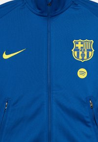 Μπλε τζάκετ με φερμουάρ, κατασκευασμένο από α παλό ύφασμα, με κίτρινο λογότυπο Nike, σήμα της Μπαρτσελόνα και έμβλημα της Spotify. Δύο πλαϊνές τσέπες με φερμουάρ.