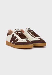 Zapatillas de ante blancas y marrones con una parte superior texturizada, punta redonda, cordones marrones y una suela de goma beige. Marca en la lengüeta.