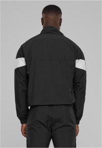 Juodas vėjo nepraleidžiantis džemperis su baltais akcentais ant rankovių. Turi stovinčią apykaklę, elastines rankoves ir sportinį dizainą.