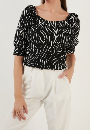 Femme portant un haut à manches bouffantes imprimé zèbre noir et blanc, rentré dans un pantalon blanc plissé taille haute, main dans la poche.
