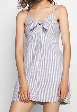 Femme portant une mini-robe sans manches à boutons, avec de fines rayures verticales grises et blanches et un nœud décoratif au niveau de la poitrine.