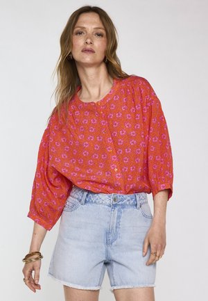 Vrouw draagt een losse rode blouse met roze bloemenprint en lichtblauwe, hooggetailleerde denimshorts met rafelige zomen, staand tegen een witte achtergrond.