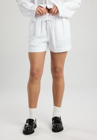 White linen shorts with an elastic waistband and drawstring detail. Smooth texture, relaxed fit, and rolled cuffs. Worn with black loafers and white socks.