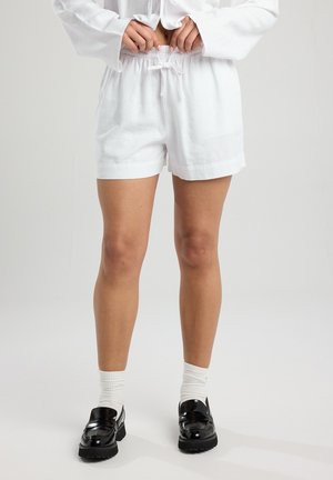 Pantaloni corti bianchi in lino con cintura elastica e dettaglio con lacci. Texture liscia, vestibilità comoda e orli arrotolati. Indossati con mocassini neri e calzini bianchi.