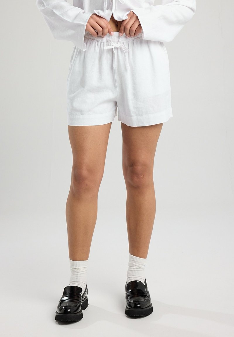 White linen shorts with an elastic waistband and drawstring detail. Smooth texture, relaxed fit, and rolled cuffs. Worn with black loafers and white socks.