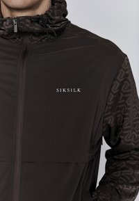 Črna jakna z visokim ovratnikom, z vzorčasto tkanino na rokavih. Sprednji žep in logo "SIKSILK" na prsih. Vodoodporen material.