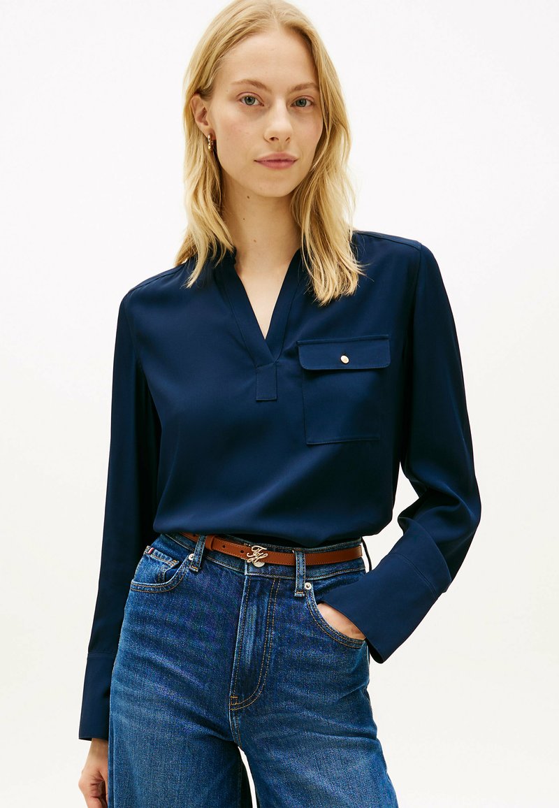 Femme blonde portant un chemisier bleu marine à manches longues avec une poche poitrine et un jean bleu taille haute avec une ceinture marron, posant avec une main dans la poche.