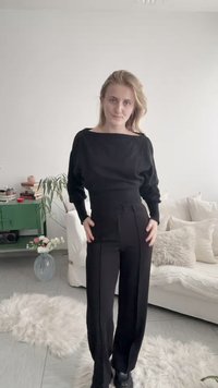 Kobieta o blond włosach, ubrana w czarną bluzkę z długim rękawem i czarne spodnie, stojąca w jasnym salonie z białą i beżową dekoracją.