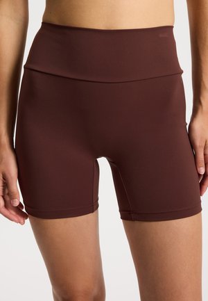 Nahaufnahme einer Person, die hoch taillierte, dunkelbraune, eng anliegende Shorts aus glattem, dehnbarem Stoff trägt.