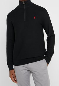 Svart stickad tröja med hög krage och fjärdedels dragkedja. Har en liten röd broderad logga på bröstet och ribbade manschetter.