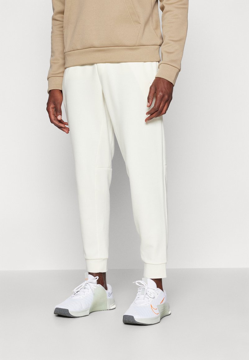 Beige hoodie with front pocket, cream jogger pants with tapered fit, white athletic shoes with gray and orange accents.