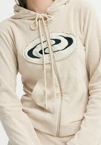 Vasaros bežinės spalvos zip-up švarkas su minkšta tekstūra, turi didelį juodą ir kreminės spalvos logotipą priekyje bei suveržiamą kapušoną.