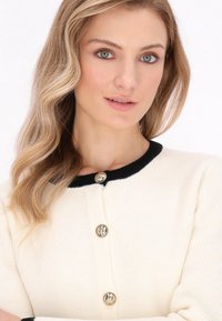 Gilet crème avec un col tricoté noir et des manches longues, arborant des boutons ronds dorés et un motif de tricot texturé.
