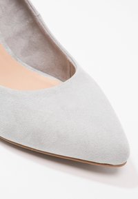 Šedá semišová bota se špičatou špičkou, hladkou texturou a nízkým střihem po stranách. Přírodní barvy stélka a minimalistický styl.