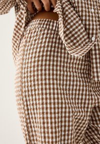 Close-up af en person iført brune og hvide ternede bukser og en matchende skjorte, med den ene hånd hvilende nær taljen.