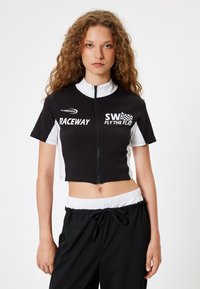 Zwart-wit gecropt shirt met een rits aan de voorkant, korte mouwen en bedrukt tekst. Beschikt over een hoge kraag en een contrasterend paneelontwerp.