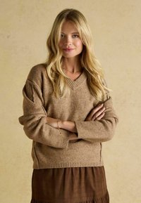 Beżowy, oversizowy sweter w serek wykonany z miękkiego dzianiny, z podwiniętymi mankietami i hemem, w połączeniu z brązową, teksturowaną spódnicą.