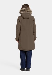 Lang, brun parka-jakke med en pelsforet hætte, der har en lige pasform og sidelommer. Båret over denimbukser og sorte støvler.