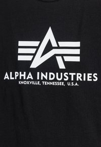 Black cotton t-shirt featuring a white logo with an A and three horizontal stripes above "ALPHA INDUSTRIES" and location text below.
