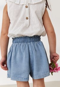Pantaloni shorts in denim azzurro chiaro con fascia elastica in vita, caratterizzati da una vestibilità comoda e una texture sottile, abbinati a un top bianco senza maniche con volant.