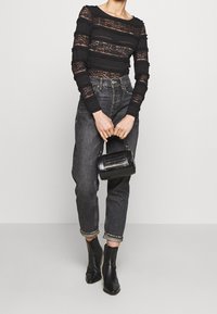 Schwarzes Spitzenoberteil mit langen Ärmeln und gestreiftem Design, hoch taillierte dunkle Jeans und schwarze Ankle Boots; hält eine kleine schwarze strukturierten Handtasche.