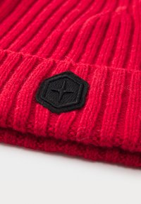 Red knitted beanie with a ribbed texture featuring a black hexagonal patch with a logo accent on the front. Soft fabric, snug fit.