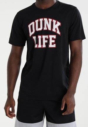Mand iført en sort t-shirt med stor hvid og rød tekst med teksten "DUNK LIFE" og sorte og grå shorts.