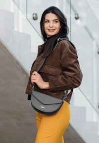 Šedá kožená taška přes rameno s zakřiveným klopou, stříbrným kováním a detailním logem, nošena s hnědou kordurovou bundou a žlutými kalhotami.