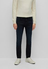 Dark blue denim jeans with a slim fit, five-pocket design, and subtle faded detail, paired with light-colored sneakers and a cream sweater.