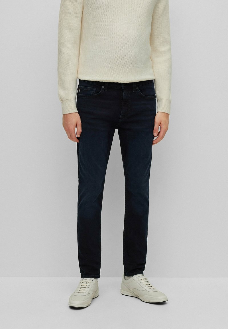 Dark blue denim jeans with a slim fit, five-pocket design, and subtle faded detail, paired with light-colored sneakers and a cream sweater.