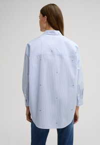 Hellblau gestreiftes Hemd mit lockerer Passform, das kleine bestickte Details aufweist. Lange Ärmel mit Knopfmanschetten und einem klassischen Kragen.