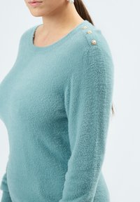 Lys blå sweater lavet af blødt, fluffy stof. Har rund halsudskæring og fire dekorative guldknapper på skulderen.