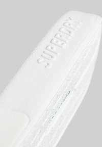 Funda de teléfono blanca texturizada con el texto en relieve "SUPERDRY", acentos lisos y un diseño intrincado a lo largo de la superficie.