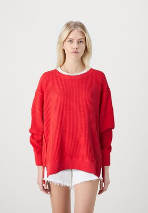 Piros, oversized pulóver texturált mintával, bordás mandzsettákkal és szegéllyel, oldalsó hasítékokkal. Fehér, réteges felső és sort nadrág fölé öltözve.