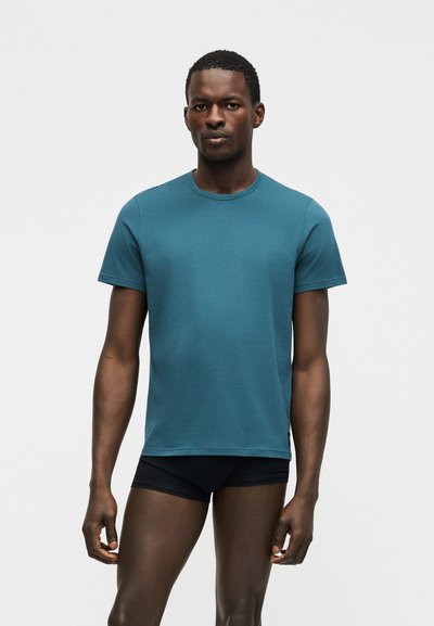 Uomo con capelli corti che indossa una maglietta a maniche corte color verde acqua e pantaloncini neri, in piedi davanti a uno sfondo grigio chiaro uniforme.